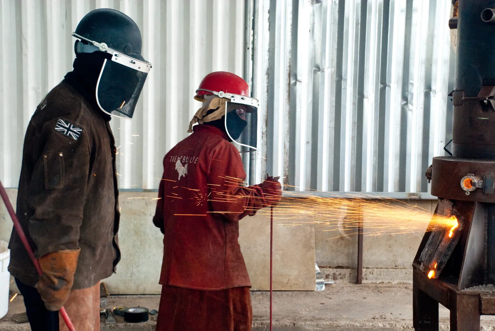 Sparks flying from the furnace as two casters in visors work beside it